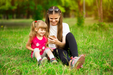 Fototapeta premium Cheerful smiling happy mother and her little daughter are sitting on green grass in summer park and using a phone for watching cartoons, a girl is indicating on the monitor at the weekend