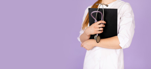 Hands of young female doctor in white uniform with phonendoscope on her neck holding black folder on purple background with space for text. Healthcare concept