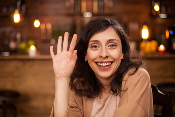 Close up portrait of beautiful young woman waving at camera in coffee shop