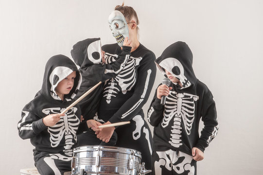 Happy friendly family of musicians in carnival costumes, boys and young mother play drum and try to sing with microphone. Black suit with image of skeletons. Classic halloween costume. Funny children