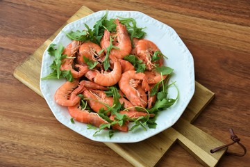 red boiled big shrimps with unpeeled heads with rucolla leaves on a white plate on a wooden board