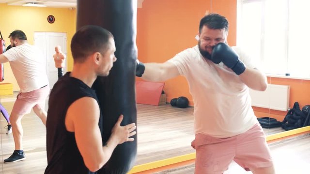 Thick man in gloves beats punching bag in gym. Individual weight loss drills for plump guy. The boxing training with personal coach. Workout with instructor in a sport hall. Private trainer for thick