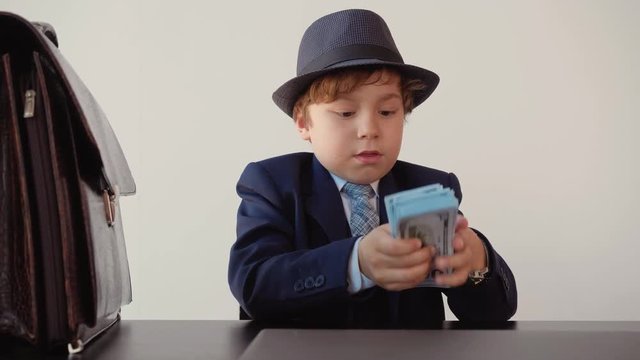 Little boy looks like a entrepreneur in suit and hat puts hundred-dollar banknotes in a stack pack on table in his office near briefcase. Business and money concept.