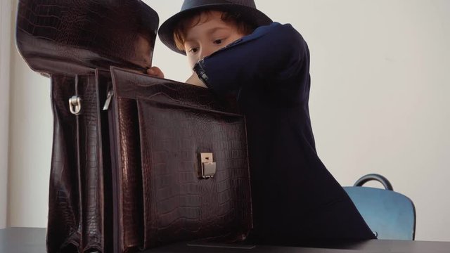 Little boy looks like a businessman in suit and hat takes a pack of hundred-dollar banknotes out of his briefcase at table in hs office. Business and money concept.
