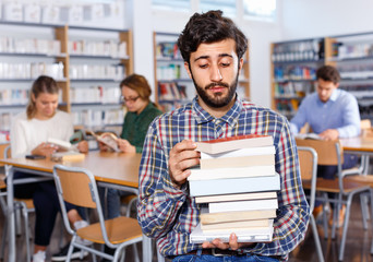 Tired man sitting with pile of books in university library on background with working fellow students