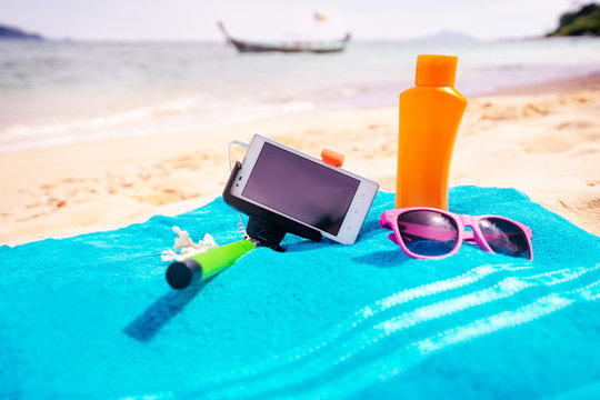 Vacation, technology and protection. Must have accessories on the sea beach. Smartphone, selfie stick, sunscreen and sunglasses.