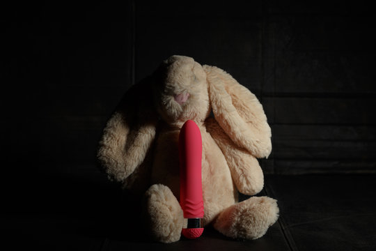 Soft toy bunny with pink dildo on a dirty dark background with hard light and shadows. Sex maniac or crazy rabbit conception.
