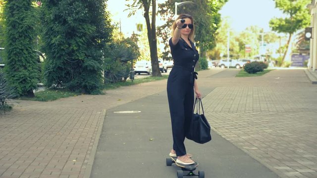 Modern business woman in a black jumpsuit in pink sneakers, sunglasses and a black bag riding on an electric board along a city street, depicting a super man, stretching one arm forward. The concept