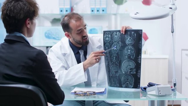 Doctor at desk in modern research facility showing X ray scans to patient. In the background - monitors displaying 3D brain simulations and DNA strings