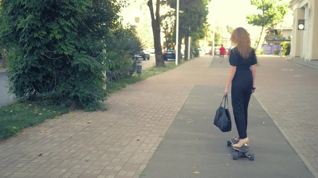A modern business woman in a black jumpsuit in beige pumps shoes, and with a black bag riding on an electric board along a city street. Concept of strange people adventures