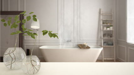 White table top or shelf with glass vase with hydroponic plant, ornament, root of plant in water, branch in vase, house plant, modern blurred bathroom background, interior design