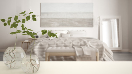 White table top or shelf with glass vase with hydroponic plant, ornament, root of plant in water, branch in vase, house plant, modern blurred bedroom in the background, interior design