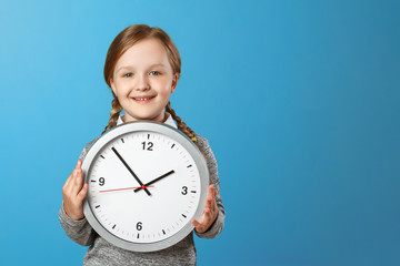 A cute little girl holding a big clock over a blue background. The concept of education, school,...