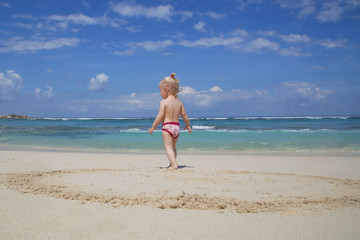 Back view of playful toddler girl on the white sandy beach looking at the horizon