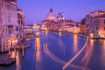 Fototapeta na wymiar Grand canal with light trails from passing boats and Basilica of Saint Mary of Health (Santa Maria della Salute) in the distance, after sunset, at night. View from the Academy bridge in Venice, Italy.
