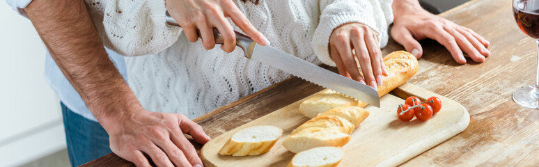 cropped view of man standing near woman during cutting bread, panoramic shot