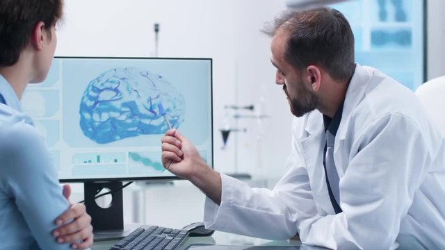 Caucasian scientist in white medical uniform explaining the intervention risks to a young female. On the screen - a 3D brain simulation