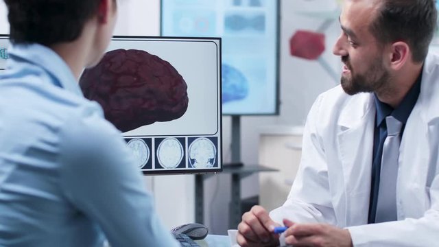Caucasian physician talking about brain damage and explaining the potential risks on a 3D simulation displayed on computer screen