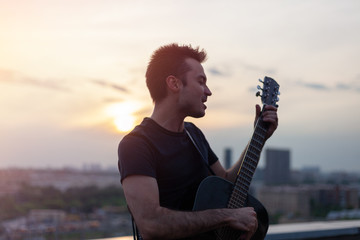 Young attractive man playing guitar and singing on the roof at sunset