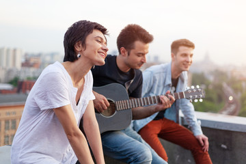 Group of friends singing songs with a guitar on the roof at sunset, summer friendship music concept