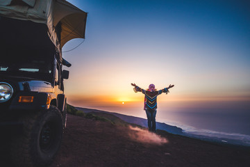 Travel and happiness concept for wanderlust people - woman with coloured warm clothes enjoy freedom and sunset near a car with tent on the roof - independence and wild traveler lifestyle female