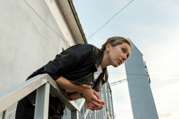Melancholic lonely young woman in black leather jacket posing on stairs with wall on background. Concept of loneliness.