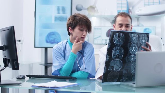 Caucasian doctor and young nurse analyzing MRI scans. In the background - modern research facility with caomputers displaying 3D brain simulations