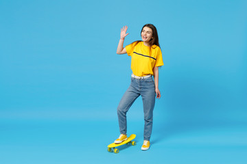 Laughing young woman looking aside, waving and greeting with hand as notices someone, standing with yellow skateboard isolated on blue wall background. People lifestyle concept. Mock up copy space.