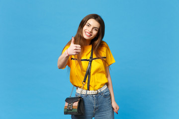 Portrait of smiling young woman in vivid casual clothes with cross body bag showing thumb up,...