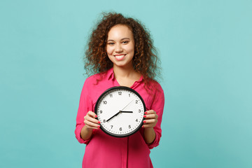 Portrait of smiling pretty african girl in pink casual clothes holding round clock isolated on blue...