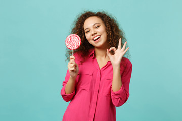 Funny african girl in casual clothes hold in hand pink round lollipop showing OK gesture isolated...