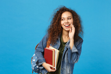 Young african american girl teen student in denim clothes, backpack hold books isolated on blue wall background studio portrait. Education in high school university college concept. Mock up copy space