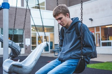 Sad teenager sitting on a swing outside of a school. He is reminiscing about when he was younger.