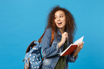 Young african american girl teen student in denim clothes, backpack hold clock isolated on blue...