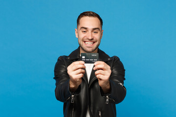 Handsome stylish young unshaven man in black leather jacket white t-shirt hold in hand credit bank card isolated on blue wall background studio portrait. People lifestyle concept. Mock up copy space.