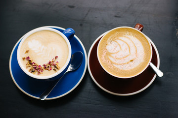 Two cups of cappuccino with latte art on black background. Beautiful foam, blue and brown ceramic cups, stylish toning, place for design and text.