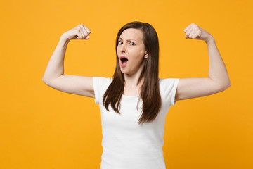 Portrait of strong young woman in white casual clothes looking camera, showing biceps, muscles isolated on bright yellow orange wall background in studio. People lifestyle concept. Mock up copy space.