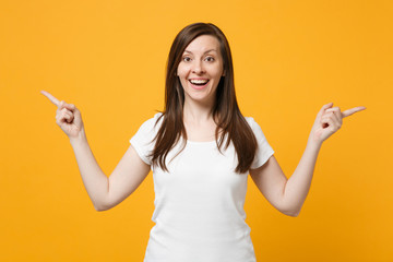 Portrait of excited young woman in white casual clothes looking camera, pointing index fingers aside isolated on yellow orange wall background in studio. People lifestyle concept. Mock up copy space.