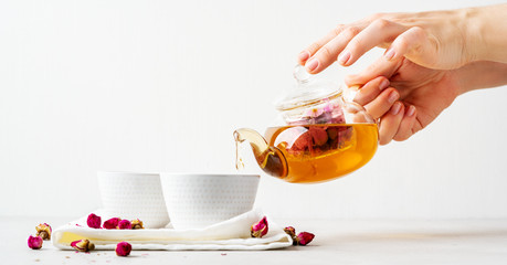 Female hands pouring tea of dry pink rose buds from transparent kettle to teacup on white...