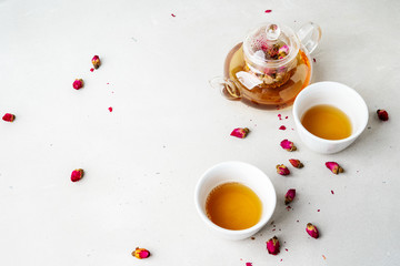 Two white cups and transparent teapot with herbal tea of dried pink roses buds over gray background with copy space. Brewing and Drinking tea.