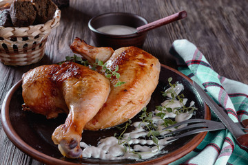 Half of baked chicken with crispy crust and spicy herbs on a ceramic plate on a wooden table next to small rye-bread buns and white sauce is ready to eat