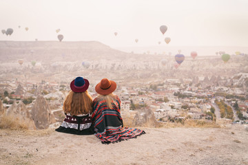 Traveling women wearing authentic boho chic style poncho, sweeter and hats looking on air ballons in sky in Cappadocia valley. Travel and wanderlust concept. Copy space background. - obrazy, fototapety, plakaty