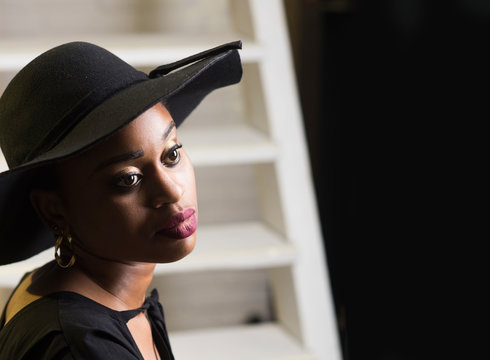 Lady on dreamy face with makeup. Lady in hat with sensual big lips. African female beauty concept. Woman with african appearance in black dress and hat looks gorgeous, interior background