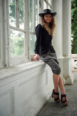 fashionable girl dressed in black shirt, hat and wide trousers posing near old white house