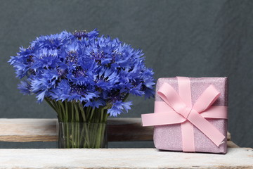 Bouquet of cornflowers in a glass vase. Next gift, packaged in a box and tied with a bow. On a gray background.