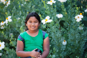 A 9 year old child or student wearing a backpack and holding a thermos standing in front of blooming flowers during a school field trip.