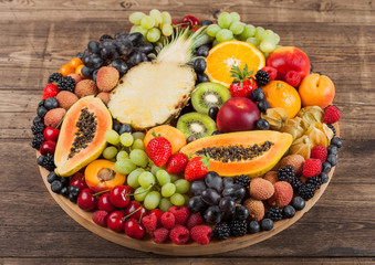 Fresh raw organic summer berries and exotic fruits in round large tray on wooden kitchen background. Papaya, grapes, nectarine, orange, raspberry, kiwi, strawberry, lychees, cherry.