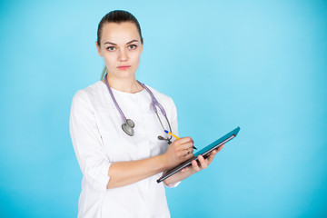 Young smiling female blonde doctor with phonendoscope on her neck and tablet on light blue background with space for text.