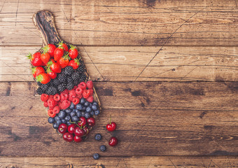 Fresh organic summer berries mix on vintage wooden chopping board on light wooden table background. Raspberries, strawberries, blueberries, blackberries and cherries. Space for text