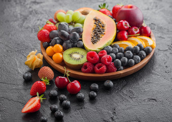 Fresh raw organic summer berries and exotic fruits in round wooden plate on black kitchen background. Papaya, grapes, nectarine, orange, raspberry, kiwi, strawberry, lychees, cherry and physalis.